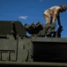 A soldier in camouflage uniform is working on top of a large military armored vehicle, likely performing maintenance or equipment checks. The vehicle is olive drab green and features visible weapon systems including what appear to be grenade launchers or smoke dischargers. The image is taken from a low angle against a clear blue sky.