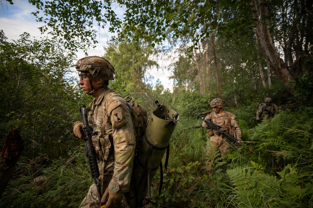 A patrol of at least three soldiers in full combat gear moves through dense woodland terrain. The lead soldier carries an assault rifle and a large cylindrical pack, with a unit patch visible on the left shoulder. The image appears to depict a military training exercise or operational patrol in a forested environment.
