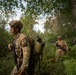 A patrol of at least three soldiers in full combat gear moves through dense woodland terrain. The lead soldier carries an assault rifle and a large cylindrical pack, with a unit patch visible on the left shoulder. The image appears to depict a military training exercise or operational patrol in a forested environment.