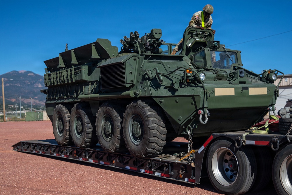 A military armored wheeled vehicle, consistent with a Stryker-type 8x8 armored personnel carrier, is loaded onto a flatbed semi-trailer. A soldier wearing camouflage uniform and a high-visibility vest is positioned on top of the vehicle, likely supervising or assisting with the loading process. The scene appears to take place at a military installation with mountains visible in the background.