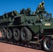 A military armored wheeled vehicle, consistent with a Stryker-type 8x8 armored personnel carrier, is loaded onto a flatbed semi-trailer. A soldier wearing camouflage uniform and a high-visibility vest is positioned on top of the vehicle, likely supervising or assisting with the loading process. The scene appears to take place at a military installation with mountains visible in the background.