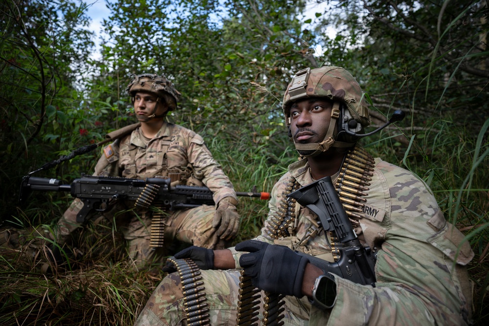 Two U.S. Army soldiers in full combat gear are positioned in a vegetated area, likely during a training exercise or field operation. Both soldiers are wearing Operational Camouflage Pattern (OCP) uniforms and helmets, and are carrying belt-fed ammunition and weapons. The foreground soldier wears a tactical headset and has 'ARMY' visible on his uniform.
