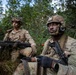 Two U.S. Army soldiers in full combat gear are positioned in a vegetated area, likely during a training exercise or field operation. Both soldiers are wearing Operational Camouflage Pattern (OCP) uniforms and helmets, and are carrying belt-fed ammunition and weapons. The foreground soldier wears a tactical headset and has 'ARMY' visible on his uniform.