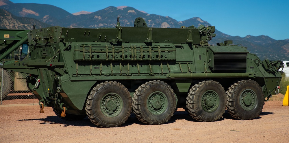 A large 8-wheeled military armored vehicle, consistent with a Stryker-family platform, is parked on a gravel/dirt surface in what appears to be a military installation. The vehicle is painted in olive drab green and features multiple external mounting rails, equipment attachments, and a remote weapon station on top. Mountains and a clear blue sky are visible in the background.