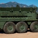 A large 8-wheeled military armored vehicle, consistent with a Stryker-family platform, is parked on a gravel/dirt surface in what appears to be a military installation. The vehicle is painted in olive drab green and features multiple external mounting rails, equipment attachments, and a remote weapon station on top. Mountains and a clear blue sky are visible in the background.