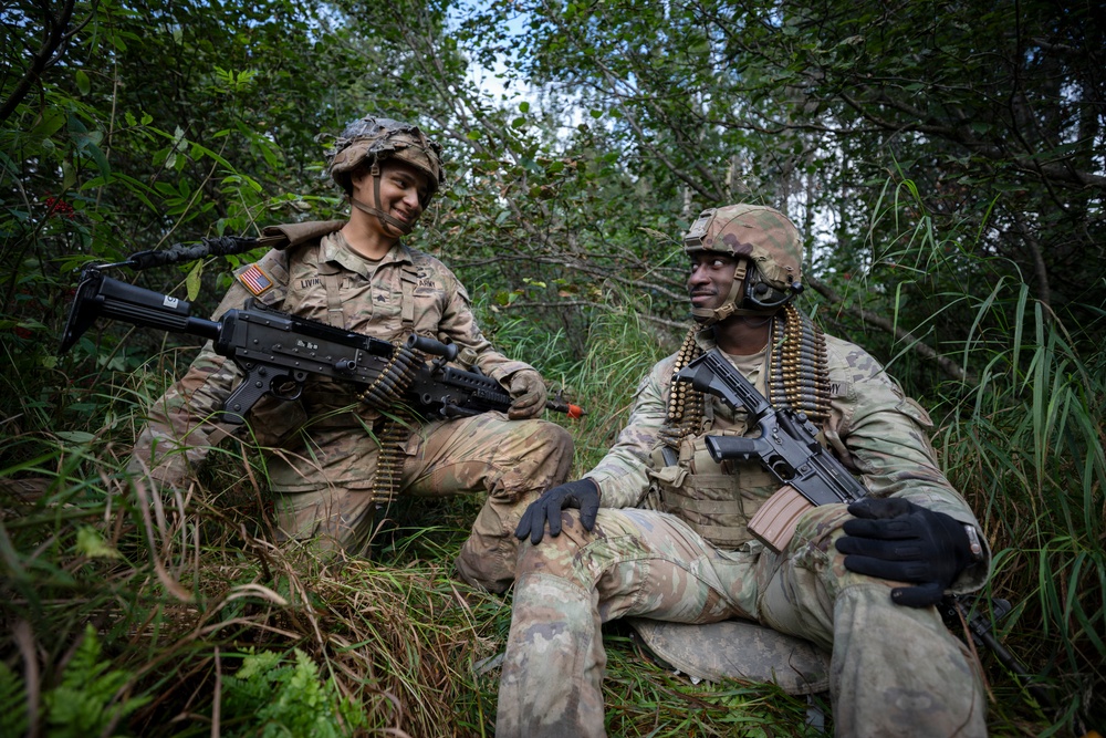 Two U.S. Army soldiers in full combat gear are positioned in a vegetated area, appearing to be on a training exercise or field operation. Both wear OCP (Operational Camouflage Pattern) uniforms with helmets and tactical equipment. They appear relaxed and are engaged in conversation while holding their weapons.