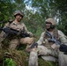 Two U.S. Army soldiers in full combat gear are positioned in a vegetated area, appearing to be on a training exercise or field operation. Both wear OCP (Operational Camouflage Pattern) uniforms with helmets and tactical equipment. They appear relaxed and are engaged in conversation while holding their weapons.