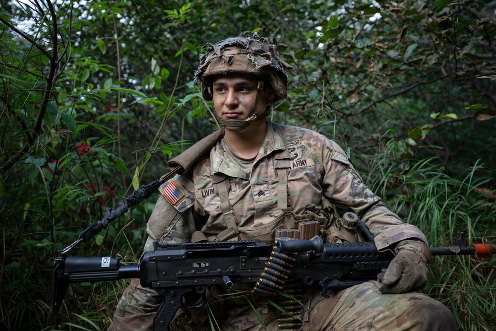 A U.S. Army soldier identified by name tape as 'LIVIN' is positioned in dense vegetation, holding a belt-fed machine gun (appears to be an M249 SAW or M240). The soldier wears Operational Camouflage Pattern (OCP) uniform with a Sergeant rank insignia, an American flag patch on the left shoulder, and a combat helmet with foliage attached. The image appears to be taken during a field training exercise.