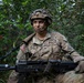 A U.S. Army soldier identified by name tape as 'LIVIN' is positioned in dense vegetation, holding a belt-fed machine gun (appears to be an M249 SAW or M240). The soldier wears Operational Camouflage Pattern (OCP) uniform with a Sergeant rank insignia, an American flag patch on the left shoulder, and a combat helmet with foliage attached. The image appears to be taken during a field training exercise.
