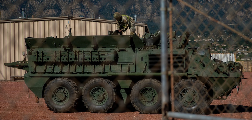 A large 8-wheeled armored military vehicle, consistent with a Stryker Infantry Carrier Vehicle, is parked on a red dirt surface within a secured compound. One soldier in camouflage uniform is visible on top of the vehicle, appearing to perform maintenance or inspection. The scene is photographed through a chain-link fence, with a mountain range and residential area visible in the background.