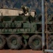 A large 8-wheeled armored military vehicle, consistent with a Stryker Infantry Carrier Vehicle, is parked on a red dirt surface within a secured compound. One soldier in camouflage uniform is visible on top of the vehicle, appearing to perform maintenance or inspection. The scene is photographed through a chain-link fence, with a mountain range and residential area visible in the background.