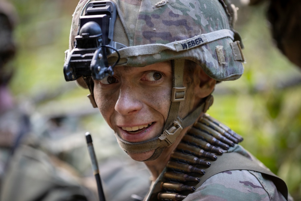 A soldier wearing MultiCam/OCP pattern camouflage gear is photographed in a close-up portrait. The soldier has face paint applied, wears a helmet with a night vision device mount, and carries a belt of ammunition across their chest. The name tape on the helmet reads 'WEBER'.