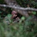 A U.S. Army soldier is photographed through dense foliage applying camouflage face paint while concealed in a wooded area. The soldier appears to be using a small mirror or compact to apply the paint, with green and dark streaks already visible on their face. The image is shot from a distance through branches and leaves, suggesting a field training or tactical exercise.