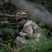 A soldier in full camouflage uniform and helmet is positioned low in dense woodland vegetation, appearing to be in a concealed observation or patrol position. The soldier is wearing face paint and holding a rifle, suggesting a military training exercise or field operation. The surrounding environment is lush green forest with fallen branches and undergrowth providing natural concealment.