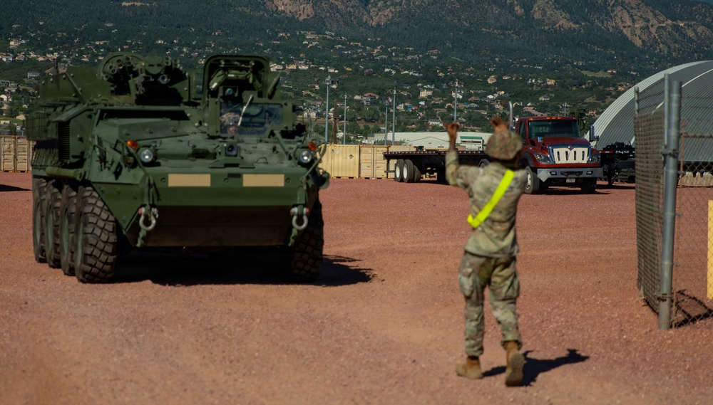 A U.S. Army soldier wearing a high-visibility yellow vest is directing a Stryker armored fighting vehicle on a military installation with a mountainous backdrop. The scene appears to be a vehicle marshaling or staging operation, with a flatbed truck visible in the background. The setting is consistent with a U.S. Army base, likely in the Colorado Springs area given the distinctive Pikes Peak region mountain profile.