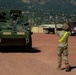A U.S. Army soldier wearing a high-visibility yellow vest is directing a Stryker armored fighting vehicle on a military installation with a mountainous backdrop. The scene appears to be a vehicle marshaling or staging operation, with a flatbed truck visible in the background. The setting is consistent with a U.S. Army base, likely in the Colorado Springs area given the distinctive Pikes Peak region mountain profile.