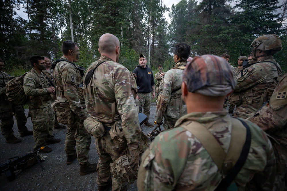 A group of military personnel in camouflage uniforms are gathered in a forested outdoor area, appearing to receive a briefing or instruction from a civilian or off-duty individual wearing a black hoodie and cap. The soldiers are equipped with tactical gear, weapons, and helmets. The setting appears to be a training exercise or operational briefing in a wooded environment.