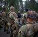 A group of military personnel in camouflage uniforms are gathered in a forested outdoor area, appearing to receive a briefing or instruction from a civilian or off-duty individual wearing a black hoodie and cap. The soldiers are equipped with tactical gear, weapons, and helmets. The setting appears to be a training exercise or operational briefing in a wooded environment.