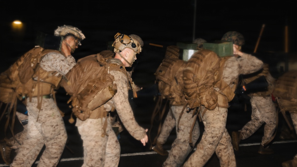 A group of military personnel, appearing to be U.S. Marines based on their desert MARPAT camouflage uniforms, are moving in formation during nighttime hours. They are heavily laden with tactical backpacks and gear, suggesting a field operation, deployment, or training exercise. The image is slightly motion-blurred, indicating movement and low-light photography conditions.