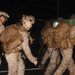A group of military personnel, appearing to be U.S. Marines based on their desert MARPAT camouflage uniforms, are moving in formation during nighttime hours. They are heavily laden with tactical backpacks and gear, suggesting a field operation, deployment, or training exercise. The image is slightly motion-blurred, indicating movement and low-light photography conditions.