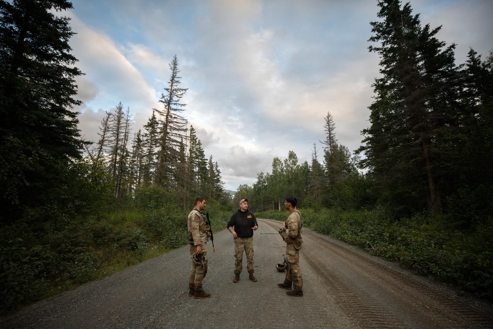 Three individuals stand on a narrow gravel road surrounded by dense coniferous forest, engaged in conversation. Two of the individuals are wearing military camouflage uniforms and carrying equipment, while the third wears a black civilian shirt and pants. The setting appears to be a remote forested area, likely during early morning or evening based on the soft, diffused lighting.