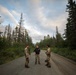 Three individuals stand on a narrow gravel road surrounded by dense coniferous forest, engaged in conversation. Two of the individuals are wearing military camouflage uniforms and carrying equipment, while the third wears a black civilian shirt and pants. The setting appears to be a remote forested area, likely during early morning or evening based on the soft, diffused lighting.