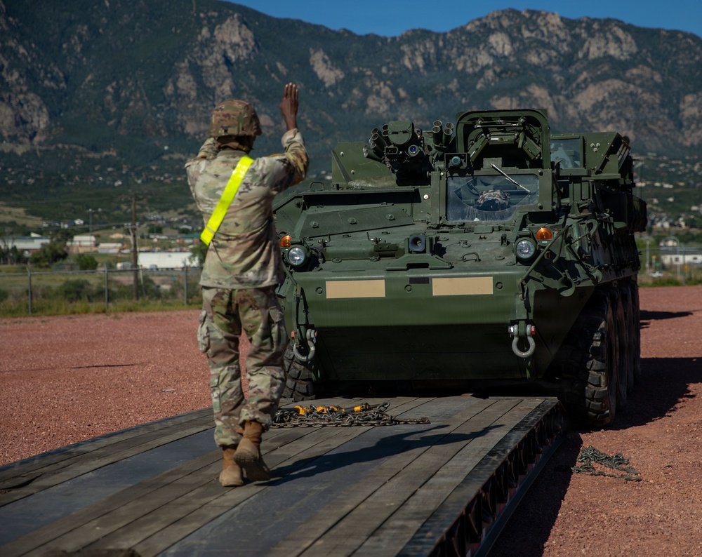 A U.S. Army soldier wearing a high-visibility safety vest is directing a Stryker armored fighting vehicle onto or off of a flatbed transport platform. The operation is taking place at what appears to be a military installation, with the Rocky Mountains visible in the background, suggesting a location such as Fort Carson, Colorado. Chains on the platform indicate tie-down equipment used to secure the vehicle during transport.