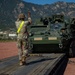 A U.S. Army soldier wearing a high-visibility safety vest is directing a Stryker armored fighting vehicle onto or off of a flatbed transport platform. The operation is taking place at what appears to be a military installation, with the Rocky Mountains visible in the background, suggesting a location such as Fort Carson, Colorado. Chains on the platform indicate tie-down equipment used to secure the vehicle during transport.