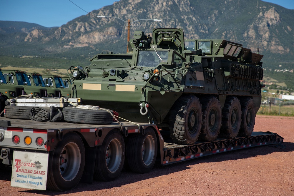 A U.S. Army Stryker armored fighting vehicle is loaded on a Tri-State Trailer Sales flatbed semi-trailer, likely being transported to or from a military installation. The setting appears to be a staging or logistics area with additional military vehicles visible in the background, set against a mountainous landscape consistent with the Colorado Front Range. The vehicle is in olive drab green livery with no visible unit markings.