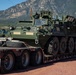 A U.S. Army Stryker armored fighting vehicle is loaded on a Tri-State Trailer Sales flatbed semi-trailer, likely being transported to or from a military installation. The setting appears to be a staging or logistics area with additional military vehicles visible in the background, set against a mountainous landscape consistent with the Colorado Front Range. The vehicle is in olive drab green livery with no visible unit markings.