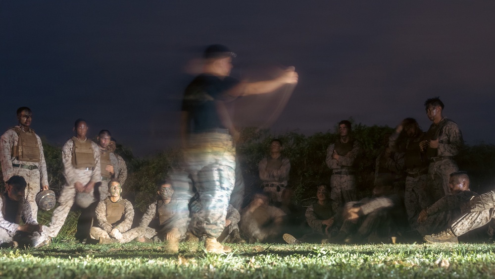 A nighttime outdoor military training or briefing session captured with a long exposure photograph. A central figure, likely an instructor or leader, appears motion-blurred due to movement during the long exposure, while surrounding personnel in military camouflage uniforms remain relatively still. The group is gathered on a grass field with vegetation visible in the background.