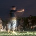 A nighttime outdoor military training or briefing session captured with a long exposure photograph. A central figure, likely an instructor or leader, appears motion-blurred due to movement during the long exposure, while surrounding personnel in military camouflage uniforms remain relatively still. The group is gathered on a grass field with vegetation visible in the background.