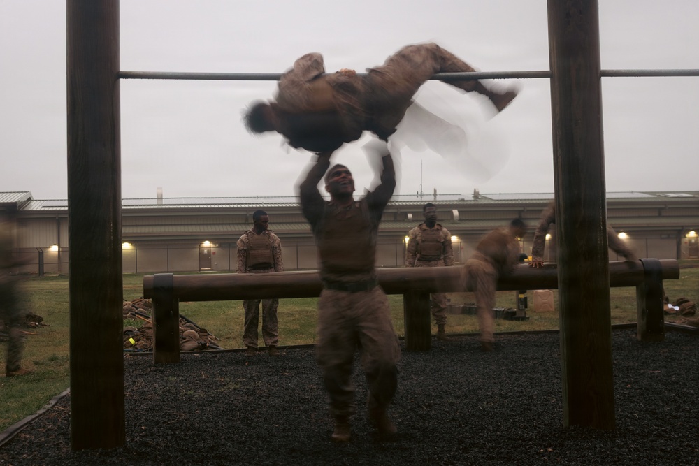 Military personnel are conducting physical training on an outdoor obstacle course during overcast, low-light conditions. The image captures motion blur of a soldier performing pull-ups or a muscle-up on a horizontal bar, with other service members visible in the background. The setting appears to be a military installation with barracks-style buildings visible in the rear.