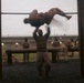 Military personnel are conducting physical training on an outdoor obstacle course during overcast, low-light conditions. The image captures motion blur of a soldier performing pull-ups or a muscle-up on a horizontal bar, with other service members visible in the background. The setting appears to be a military installation with barracks-style buildings visible in the rear.