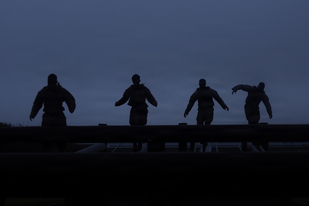 Four silhouetted figures are visible against a dark blue twilight sky, positioned along what appears to be a raised horizontal structure such as a wall, beam, or barrier. The figures appear to be wearing tactical or military-style gear with backpacks or vests. The image is taken at dusk or dawn, creating a dramatic low-light silhouette effect.
