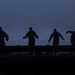 Four silhouetted figures are visible against a dark blue twilight sky, positioned along what appears to be a raised horizontal structure such as a wall, beam, or barrier. The figures appear to be wearing tactical or military-style gear with backpacks or vests. The image is taken at dusk or dawn, creating a dramatic low-light silhouette effect.