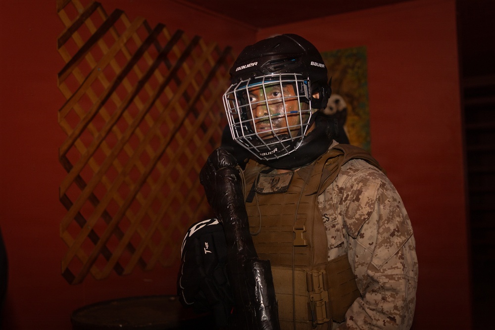 A person wearing U.S. Marine Corps digital camouflage (MARPAT) uniform with a tactical vest is equipped with a Bauer hockey helmet and cage face guard, along with a hockey glove and stick. The individual appears to have face paint applied, suggesting a training or role-playing exercise scenario. The setting appears to be an indoor environment with red walls and a lattice-style wooden panel in the background.