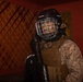 A person wearing U.S. Marine Corps digital camouflage (MARPAT) uniform with a tactical vest is equipped with a Bauer hockey helmet and cage face guard, along with a hockey glove and stick. The individual appears to have face paint applied, suggesting a training or role-playing exercise scenario. The setting appears to be an indoor environment with red walls and a lattice-style wooden panel in the background.