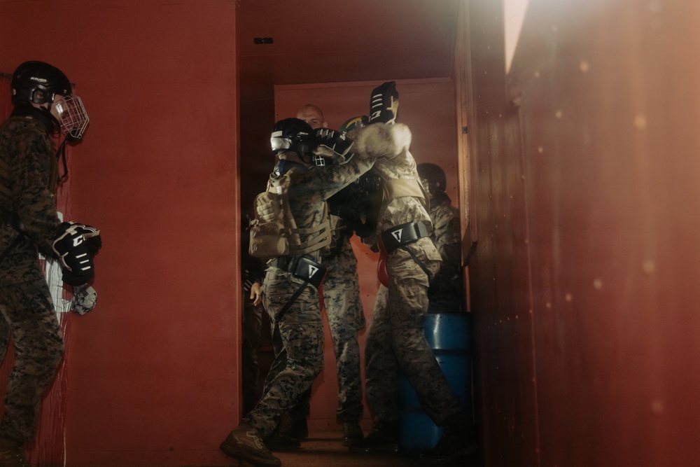 The image depicts multiple individuals in military camouflage uniforms and protective gear, including helmets with face guards and padded gloves, engaged in what appears to be a close-quarters combat training exercise. They are clustered in a doorway of a red-walled structure, with some physical grappling or restraint activity visible. The scene is lit with a warm reddish haze, suggesting a controlled training environment.