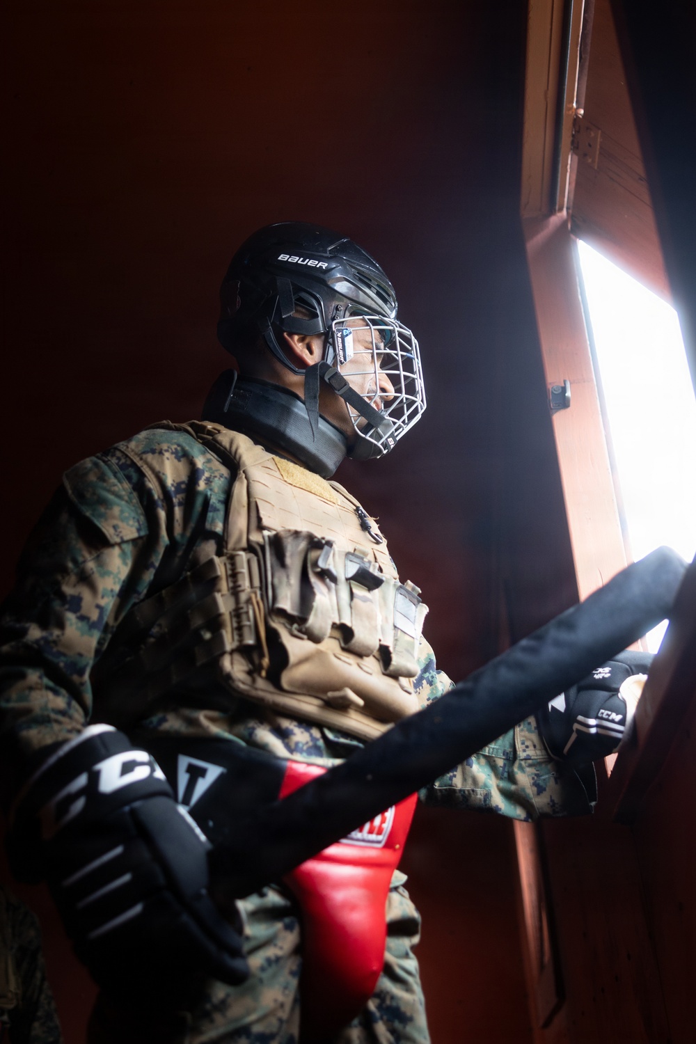 A person wearing U.S. Marine Corps MARPAT camouflage uniform is equipped with a combination of military and sports protective gear. They are wearing a Bauer hockey helmet with a wire cage face guard, a tactical plate carrier vest, CCM hockey gloves, and are holding what appears to be a padded training implement. This appears to be a military close-quarters combat or martial arts training scenario using improvised protective equipment.