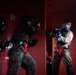 The image shows military personnel, likely U.S. Marines based on their distinctive MARPAT camouflage uniforms, engaged in close-quarters combat training inside a confined red-walled structure. The participants are wearing full protective gear including helmets with face cages, boxing/sparring gloves, and groin protectors, indicating a controlled combatives training exercise. The scene captures active sparring or fighting drills in what appears to be a military training facility.