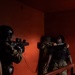 The image depicts what appears to be a military close-quarters combat training exercise in a red-walled indoor structure. Three individuals in military camouflage uniforms and protective gear are engaged in a simulated hostage or detainee scenario on a stairwell landing. One person aims a training weapon while two others simulate a grappling or restraint situation.