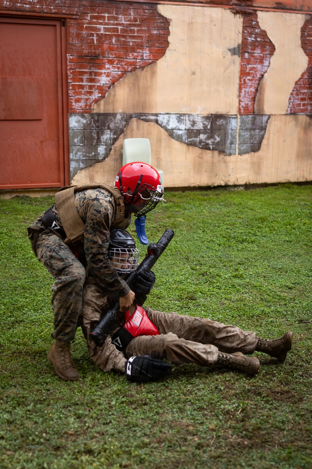 Two individuals in U.S. Marine Corps camouflage uniforms are engaged in a close-combat training exercise on a grass field. One Marine wearing a red helmet with a face cage is in a dominant ground position over another Marine who is on their back. Both are equipped with protective gear including helmets, face guards, and padded equipment, indicating a controlled martial arts or combatives training drill.