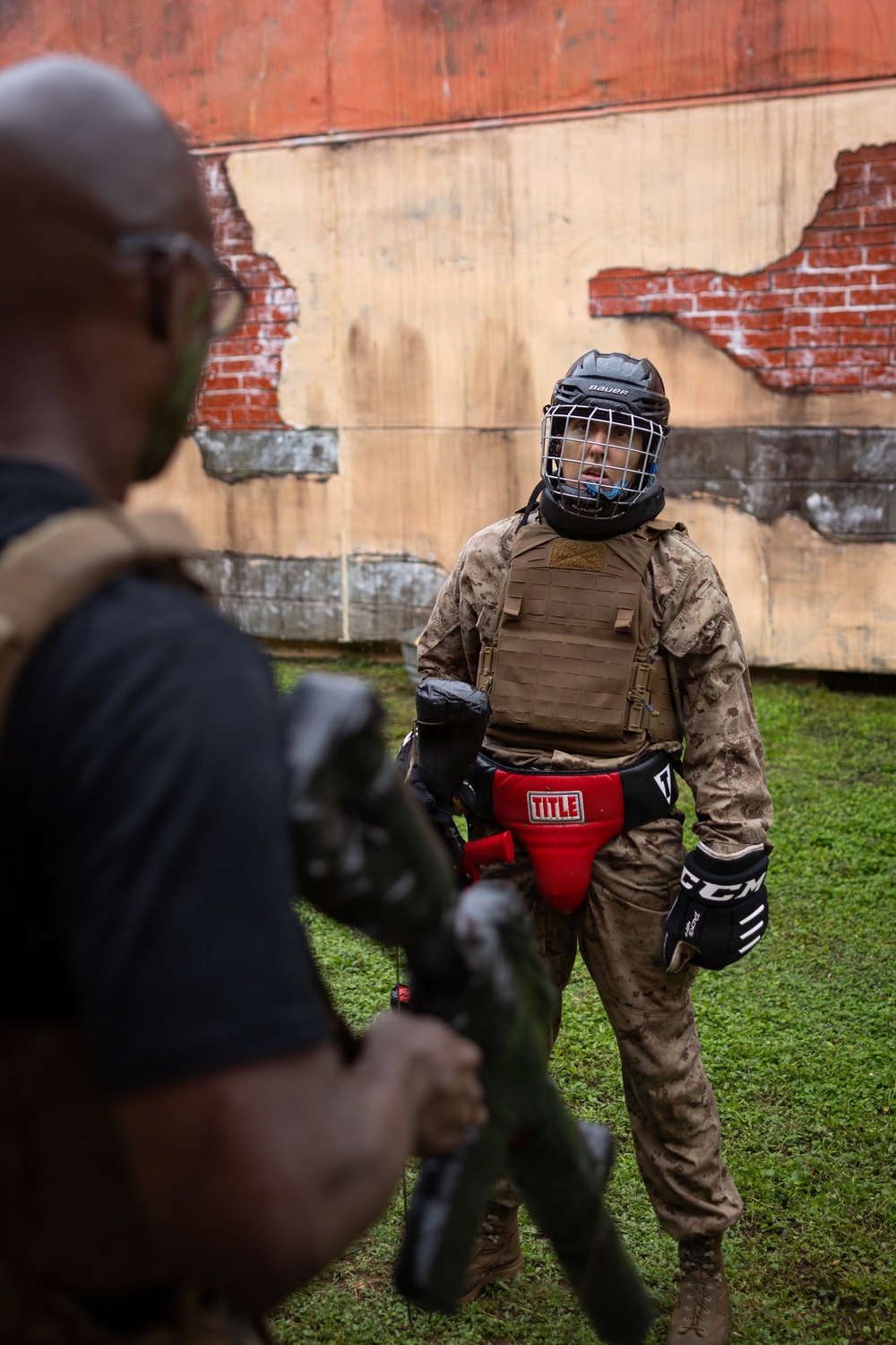 A person in military camouflage uniform is wearing full protective combat training gear, including a Bauer hockey-style helmet with cage face guard, a tactical plate carrier vest, red TITLE groin protector, and CCM hockey gloves. They appear to be engaged in a close-quarters combat or martial arts training exercise outdoors. A second individual in the foreground, partially out of focus, appears to be an instructor or opponent holding training equipment.