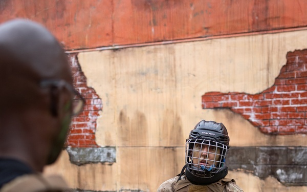 A person in military camouflage uniform is wearing full protective combat training gear, including a Bauer hockey-style helmet with cage face guard, a tactical plate carrier vest, red TITLE groin protector, and CCM hockey gloves. They appear to be engaged in a close-quarters combat or martial arts training exercise outdoors. A second individual in the foreground, partially out of focus, appears to be an instructor or opponent holding training equipment.