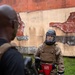 A person in military camouflage uniform is wearing full protective combat training gear, including a Bauer hockey-style helmet with cage face guard, a tactical plate carrier vest, red TITLE groin protector, and CCM hockey gloves. They appear to be engaged in a close-quarters combat or martial arts training exercise outdoors. A second individual in the foreground, partially out of focus, appears to be an instructor or opponent holding training equipment.
