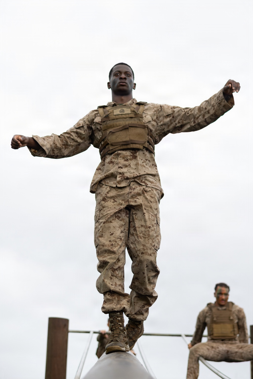 A military service member in desert digital camouflage (MARPAT) uniform and tactical vest is balancing on a narrow elevated beam or log with arms outstretched, appearing to navigate an obstacle course. A second service member in similar uniform is visible in the background to the right, seated on a platform. The image is taken from a low angle looking upward against an overcast sky.