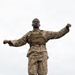 A military service member in desert digital camouflage (MARPAT) uniform and tactical vest is balancing on a narrow elevated beam or log with arms outstretched, appearing to navigate an obstacle course. A second service member in similar uniform is visible in the background to the right, seated on a platform. The image is taken from a low angle looking upward against an overcast sky.