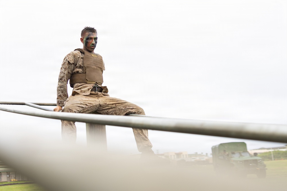 A military service member wearing camouflage utilities, a plate carrier vest, and face paint in green and brown tones is perched atop a metal structure, looking directly at the camera. The low-angle shot creates a dramatic perspective with metal rails in the foreground and a hazy, overcast sky in the background. A military vehicle is partially visible in the lower right background.