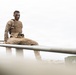 A military service member wearing camouflage utilities, a plate carrier vest, and face paint in green and brown tones is perched atop a metal structure, looking directly at the camera. The low-angle shot creates a dramatic perspective with metal rails in the foreground and a hazy, overcast sky in the background. A military vehicle is partially visible in the lower right background.